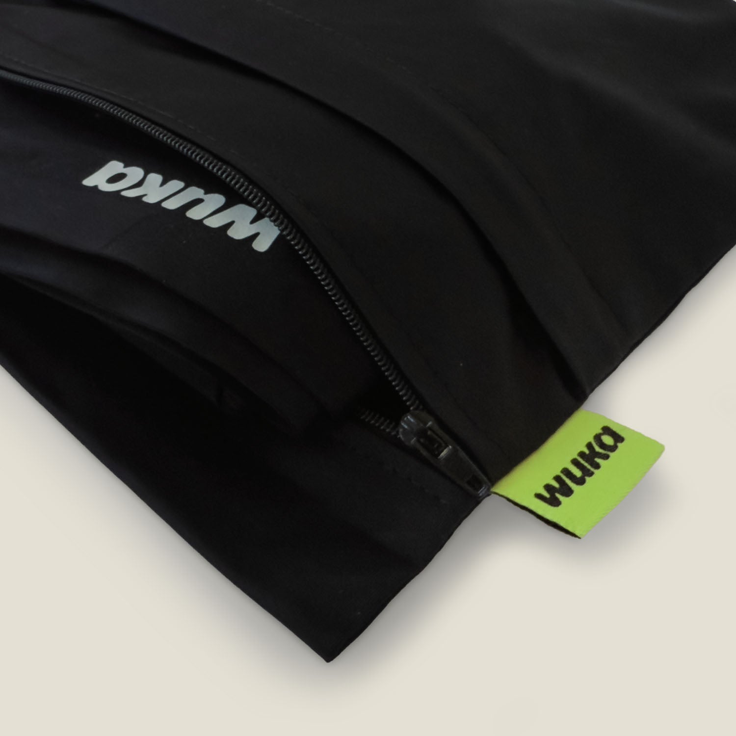 Close-up view of the WUKA Two Pocket Changing Bag showing zip detail and red branded tag, with folded WUKA pants partially tucked inside for convenient access.