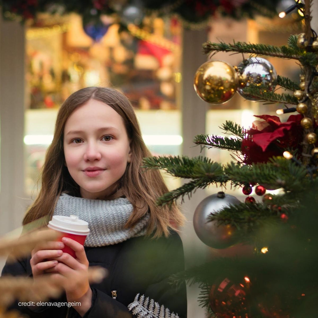 Young girl standing near a decorated Christmas tree, holding a red cup and wearing a scarf - capturing a quiet festive moment outdoors.
