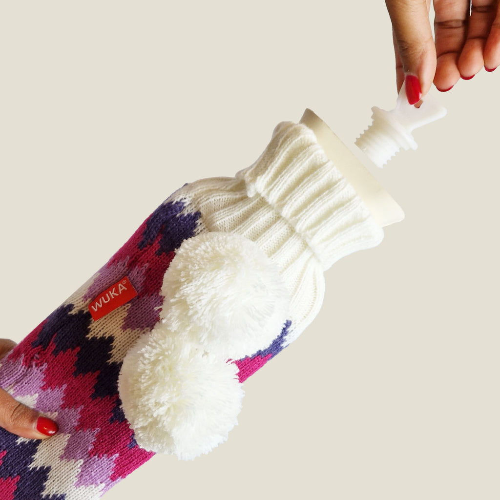 Close-up of WUKA long hot water bottle being opened, showing white screw cap and textured knit cover with pom poms.